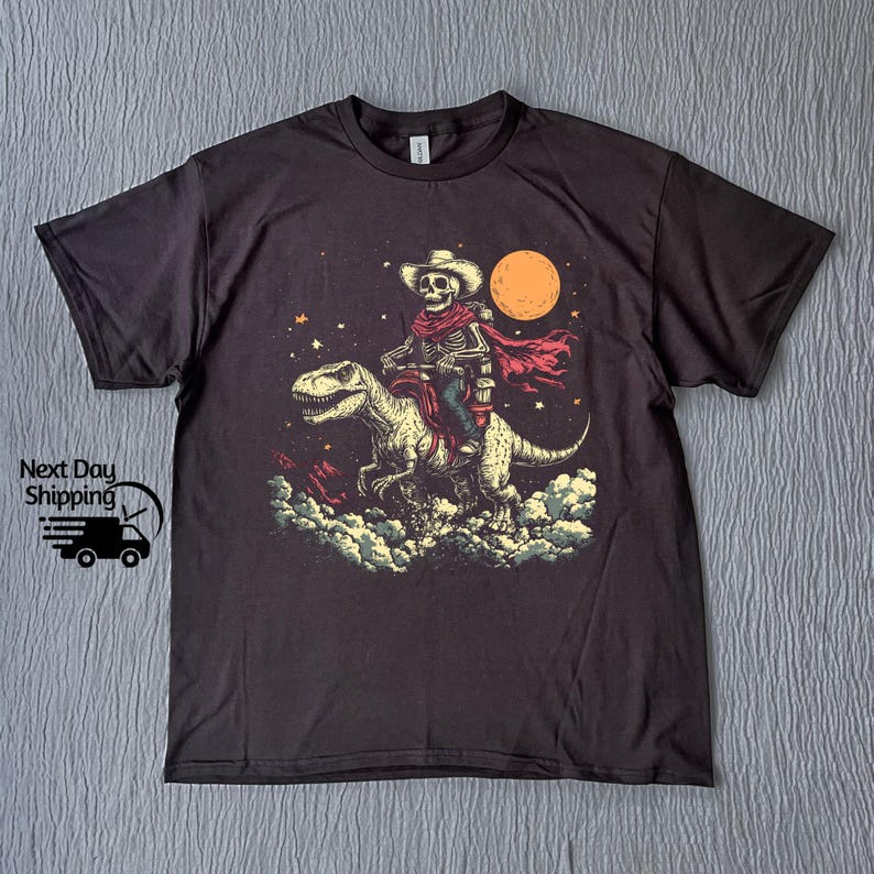 May include: Dark grey t-shirt with a graphic of a skeleton cowboy riding a dinosaur under a full moon. The cowboy wears a hat and red scarf. The graphic is in beige, red, and blue.