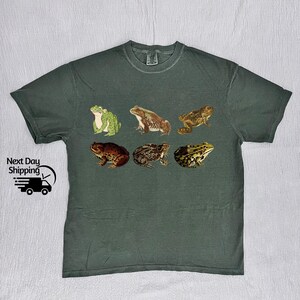 May include: A dark green t-shirt with a vintage illustration of six frogs in different poses. The frogs are in shades of green, brown, and tan.