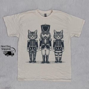 Puede incluir: Una camiseta beige con un estampado gráfico de tres gatos vestidos de cascanueces. Los gatos llevan uniformes a rayas blancas y negras con detalles rojos. El gato del medio es un soldado cascanueces con un sombrero rojo y negro y un bigote.