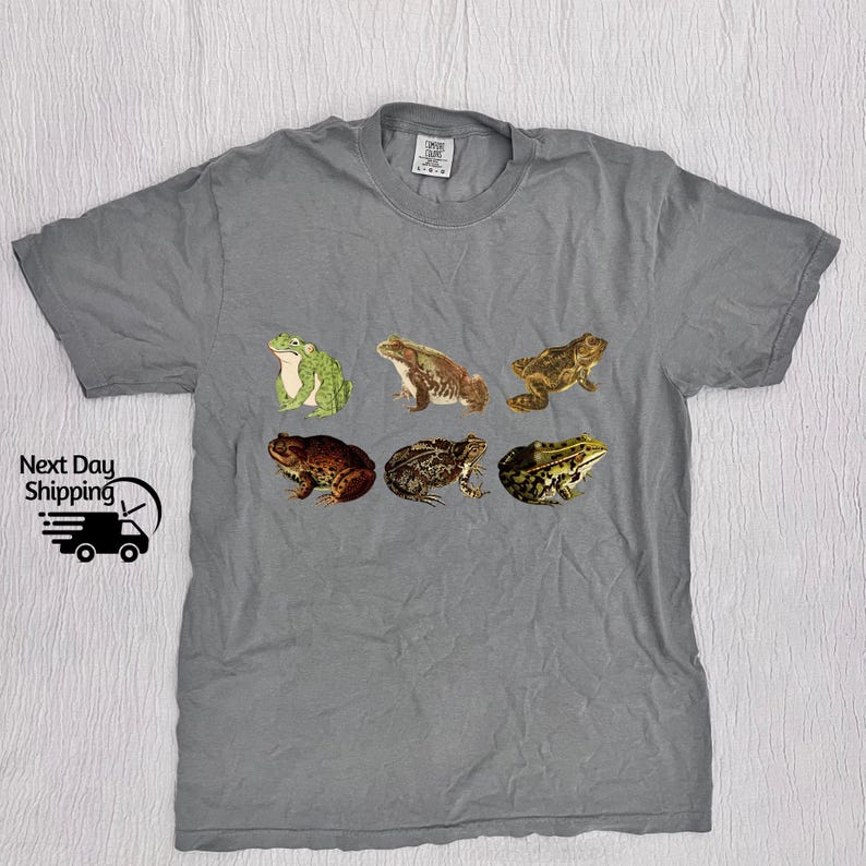 May include: Gray t-shirt with a print of six different frogs in various poses. The frogs are brown, green, and tan.