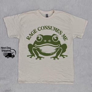 May include: A beige t-shirt with a green frog graphic and the text "Rage Consumes Me".