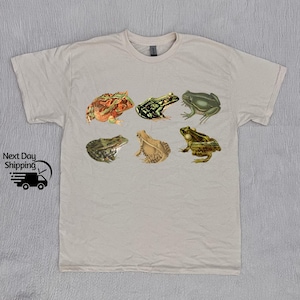Puede incluir: Una camiseta beige claro con un estampado gr&aacute;fico de seis ranas diferentes en varios colores y poses. Las ranas est&aacute;n todas mirando hacia adelante y tienen diferentes expresiones.