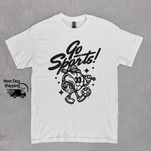 May include: White t-shirt with a black and white graphic of a cartoon character holding a baseball bat and a soccer ball. The text "Go Sports!" is written in a stylized font above the character.