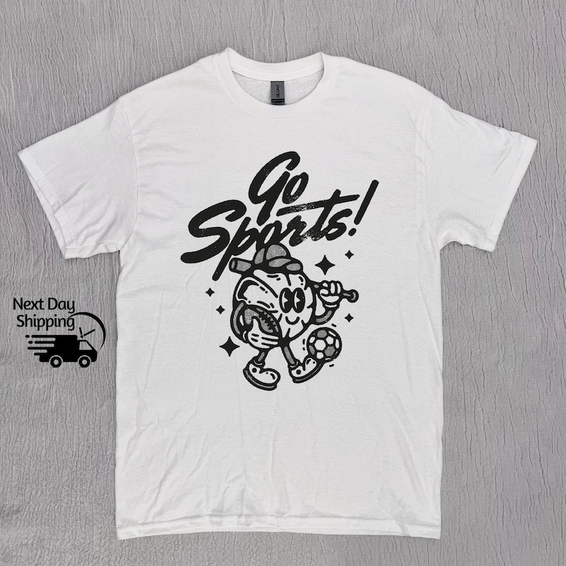 May include: White t-shirt with a black and white graphic of a cartoon character holding a baseball bat and a soccer ball. The text "Go Sports!" is written in a stylized font above the character.