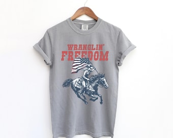 May include: A gray t-shirt with the words "Wranglin' Freedom" in red block letters. Below the text is a blue illustration of a person riding a horse while holding an American flag. The shirt is hanging on a wooden hanger.