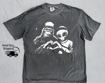 Puede incluir: Camiseta gris oscuro con un gráfico blanco de un Bigfoot y un extraterrestre formando un corazón con sus manos. El Bigfoot lleva una gorra y el extraterrestre tiene grandes ojos ovalados. Una pequeña estrella es visible encima de ellos.