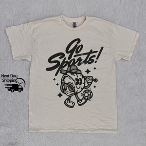May include: A beige t-shirt with a black graphic of a cartoon character playing sports. The character is holding a baseball bat and a soccer ball. The text "Go Sports!" is written above the character.
