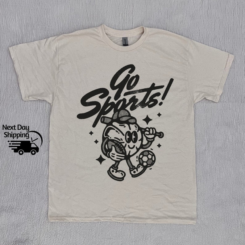 May include: A beige t-shirt with a black graphic of a cartoon character playing sports. The character is holding a baseball bat and a soccer ball. The text "Go Sports!" is written above the character.