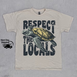 May include: A beige t-shirt with a distressed black graphic of a sea turtle and the text "Respect the Locals".