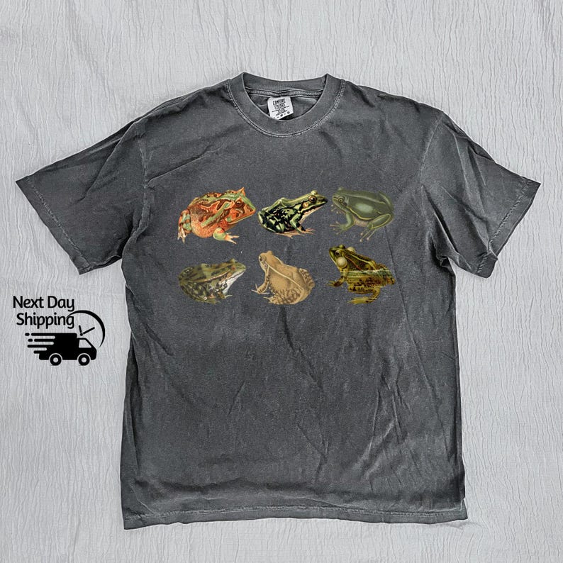 Puede incluir: Una camiseta gris oscuro con un estampado gr&aacute;fico de seis ranas diferentes. Las ranas est&aacute;n en varias poses y colores, incluyendo verde, marr&oacute;n y naranja.
