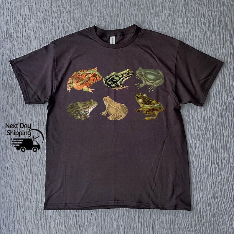Puede incluir: Una camiseta gris oscuro con una impresi&oacute;n de seis ranas diferentes en varios colores y poses.