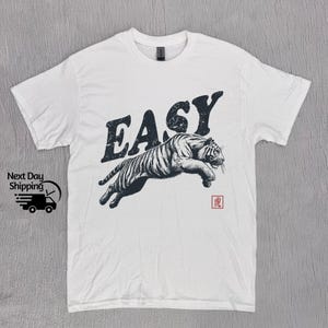 K&ouml;nnte beinhalten: Wei&szlig;es T-Shirt mit einem schwarz-wei&szlig;en Tiger-Motiv und dem Wort "EASY" in gro&szlig;en, fetten Buchstaben. Der Tiger springt und das Wort "EASY" befindet sich dar&uuml;ber. In der rechten unteren Ecke befindet sich ein kleines rotes Quadrat mit einem japanischen Schriftzeichen.