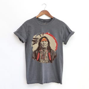 Peut inclure: Un t-shirt gris anthracite avec une illustration vintage d'un homme amérindien portant une coiffe et des vêtements traditionnels.