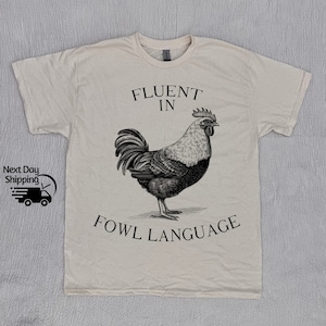 May include: A beige t-shirt with a black and white illustration of a rooster and the text "Fluent in Fowl Language".