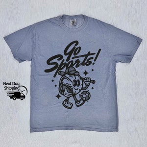 May include: A light blue t-shirt with a black graphic of a cartoon character holding a baseball bat and a soccer ball. The text "Go Sports!" is printed in black above the character.
