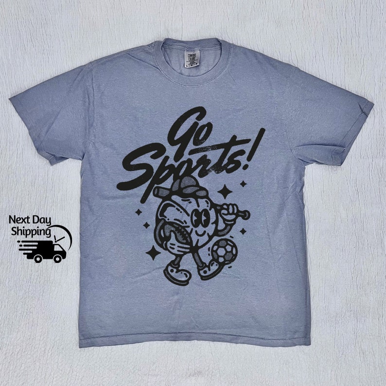 May include: A light blue t-shirt with a black graphic of a cartoon character holding a baseball bat and a soccer ball. The text "Go Sports!" is printed in black above the character.