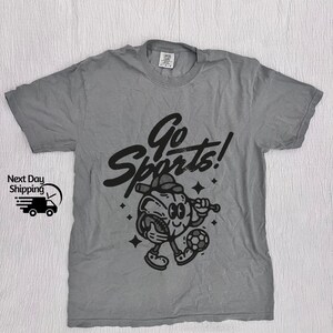 May include: A gray t-shirt with a black graphic of a cartoon character playing sports. The character is holding a baseball bat and a football. The text "Go Sports!" is written above the character.