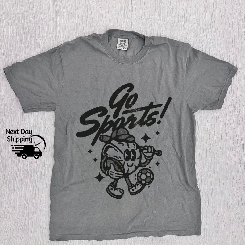 May include: A gray t-shirt with a black graphic of a cartoon character playing sports. The character is holding a baseball bat and a football. The text "Go Sports!" is written above the character.