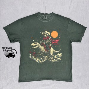 May include: Dark green t-shirt with a graphic of a skeleton cowboy riding a dinosaur under a full moon. The cowboy wears a hat and red scarf. The graphic includes stars and clouds. The shirt has the text "Next Day Shipping" and a graphic of a truck.