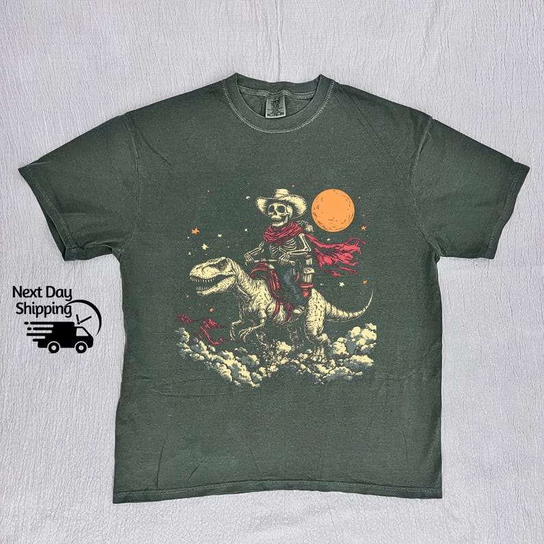 May include: Dark green t-shirt with a graphic of a skeleton cowboy riding a dinosaur under a full moon. The cowboy wears a hat and red scarf. The graphic includes stars and clouds. The shirt has the text "Next Day Shipping" and a graphic of a truck.