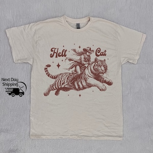 Könnte beinhalten: Ein beiges T-Shirt mit einem braunen Aufdruck einer Frau, die auf einem Tiger reitet. Der Aufdruck enthält den Text "Hell Cat" und Sterne.