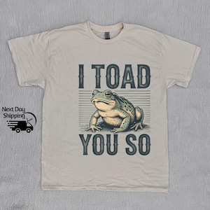 May include: Cream-coloured t-shirt with a detailed illustration of a toad and the text "I TOAD YOU SO". The toad is green and brown, set against a striped background. The design has a vintage style.