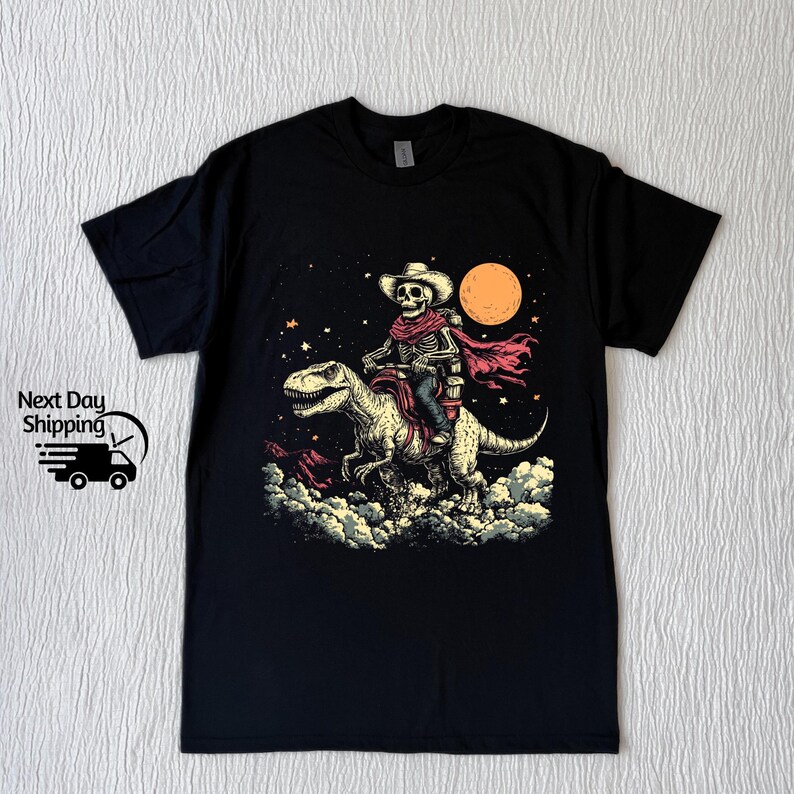May include: Black t-shirt with a graphic of a skeleton cowboy riding a dinosaur under a night sky with a full moon. The cowboy wears a hat and scarf. The dinosaur is light green and cream.
