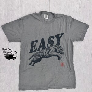 K&ouml;nnte beinhalten: Graues T-Shirt mit einem schwarz-wei&szlig;en Tiger-Motiv und dem Wort "EASY" in schwarzen Buchstaben. Der Tiger springt und das Wort "EASY" befindet sich dar&uuml;ber. Unter dem Tiger befindet sich ein kleines rotes Symbol.