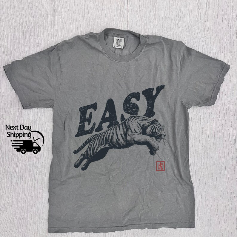 K&ouml;nnte beinhalten: Graues T-Shirt mit einem schwarz-wei&szlig;en Tiger-Motiv und dem Wort "EASY" in schwarzen Buchstaben. Der Tiger springt und das Wort "EASY" befindet sich dar&uuml;ber. Unter dem Tiger befindet sich ein kleines rotes Symbol.
