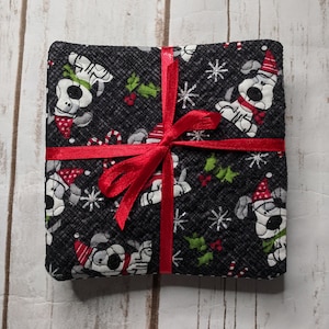 May include: A set of four black fabric coasters with a red ribbon tied around them. The coasters feature a repeating pattern of white dogs wearing Santa hats and red scarves, snowflakes, holly, and candy canes.
