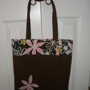 Pode incluir: Uma bolsa tote marrom com estampa floral em rosa, amarelo e azul. A bolsa tem um forro de tecido marrom e uma única flor rosa grande na frente.