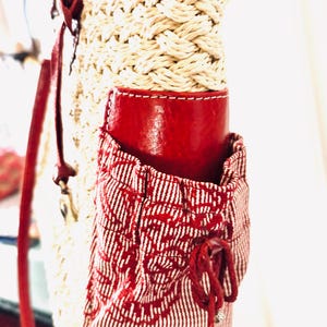 May include: A woven straw bag with a red leather accent and a red and white patterned pocket. The bag has a red leather strap and a drawstring closure on the pocket. The bag is a stylish accessory.