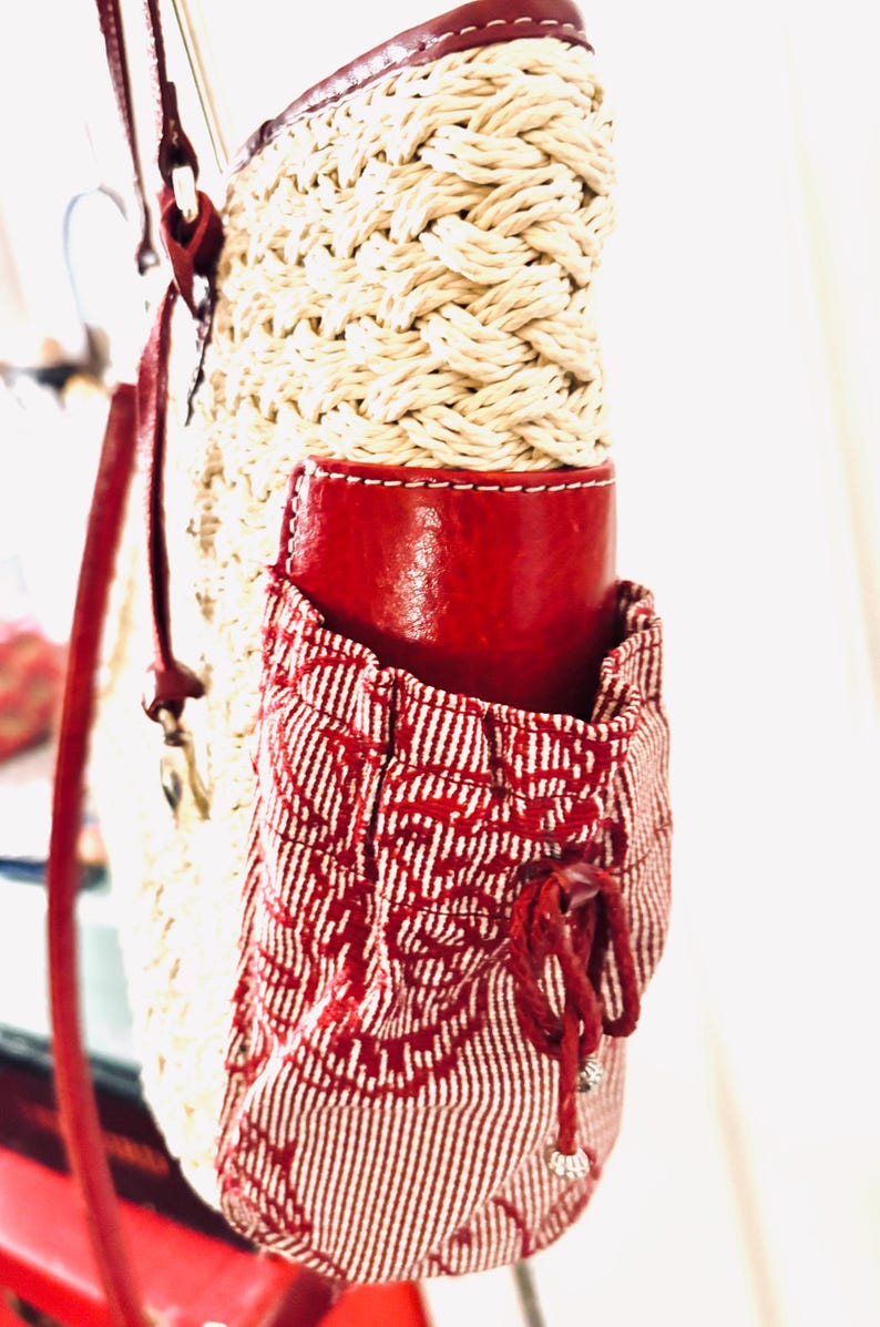 May include: A woven straw bag with a red leather accent and a red and white patterned pocket. The bag has a red leather strap and a drawstring closure on the pocket. The bag is a stylish accessory.