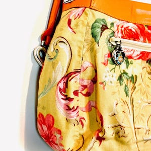 以下が含まれることがあります： 黄色い背景に赤とピンクの花柄のハンドバッグ。茶色の革のストラップとトリム、銀色のハート型のジッパープルが付いています。バッグの幅は約30cmです。