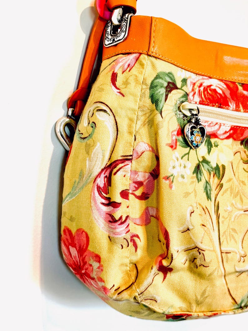 以下が含まれることがあります： 黄色い背景に赤とピンクの花柄のハンドバッグ。茶色の革のストラップとトリム、銀色のハート型のジッパープルが付いています。バッグの幅は約30cmです。