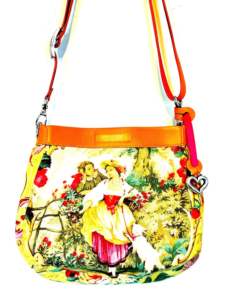 以下が含まれることがあります： カラフルな牧歌的なシーンのプリントが施された黄色のクロスボディバッグ。バッグは、赤とオレンジのストラップ、ハート型のチャーム、オレンジ色のレザートリムが特徴です。プリントは、犬と一緒に庭にいる人物を描いています。