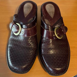 May include: A pair of brown leather clogs with a buckle detail. The clogs have a woven pattern and a slightly raised heel.