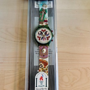May include: A Swatch watch in its original packaging. The watch features a green band with colorful designs and a round face with multiple images. The watch is an official timekeeper of the 1996 Olympic Games in Atlanta.