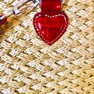 May include: Close-up of a woven straw bag with a red leather heart-shaped tag. The tag is stitched with white thread and attached to a red strap. The bag's weave is a light beige colour, and the strap has silver hardware.