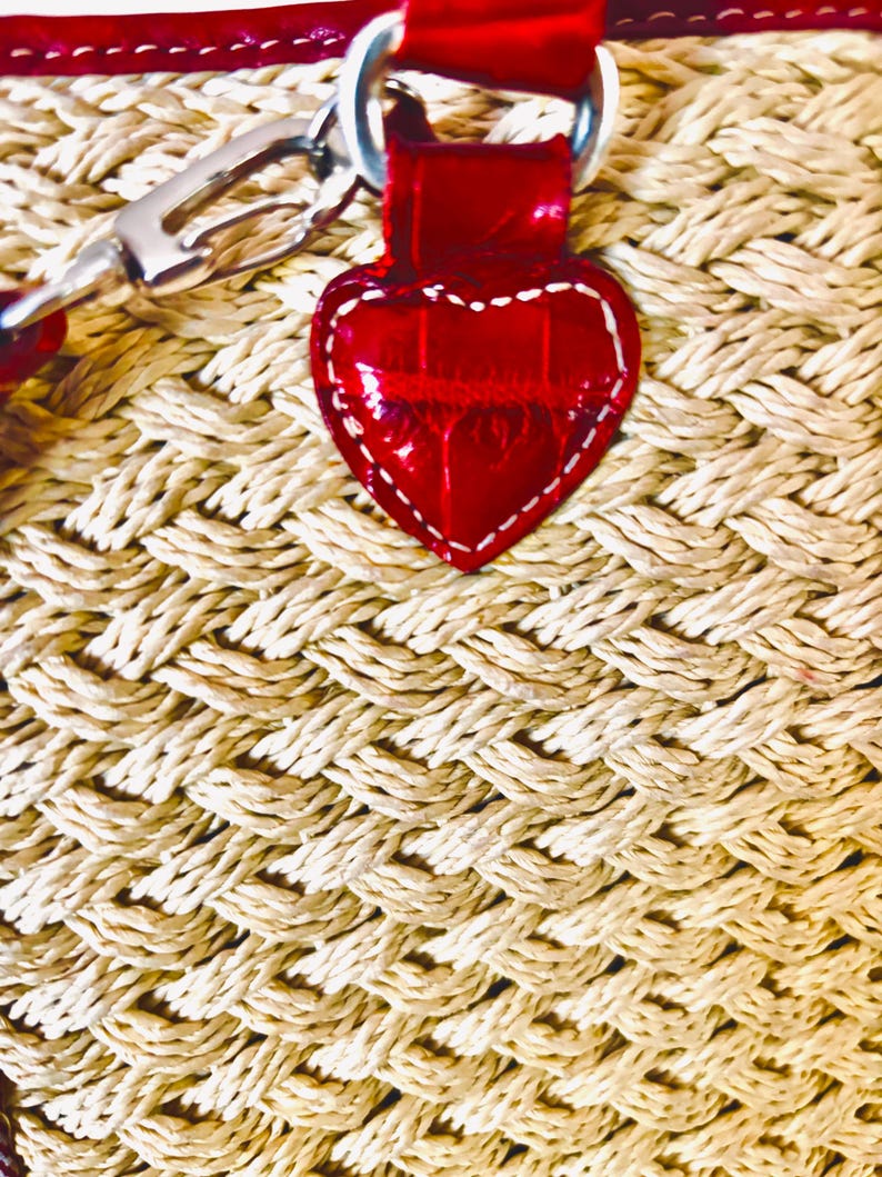 May include: Close-up of a woven straw bag with a red leather heart-shaped tag. The tag is stitched with white thread and attached to a red strap. The bag's weave is a light beige colour, and the strap has silver hardware.