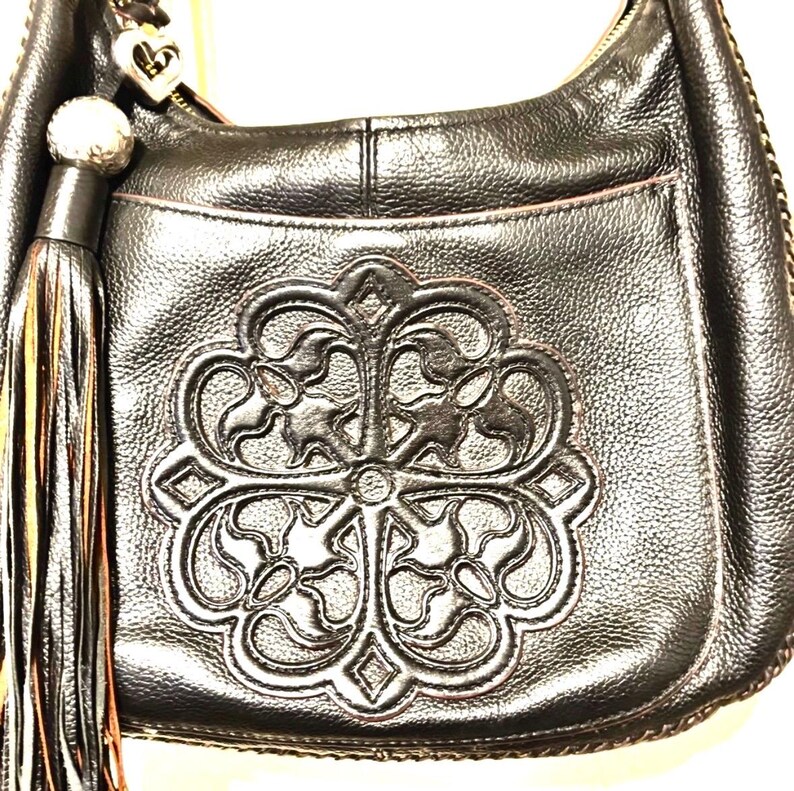 May include: Black leather shoulder bag with a decorative embossed floral design on the front. The bag features a tassel, a silver-tone zipper, and a heart-shaped charm. The bag has a textured finish and a stitched trim.