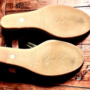 May include: Pair of wedge sandals with light brown soles and a stitched edge. The soles have a textured surface with a signature and the words "McCartney" and "Made in Italy" printed on them. The sandals are on a wooden surface.