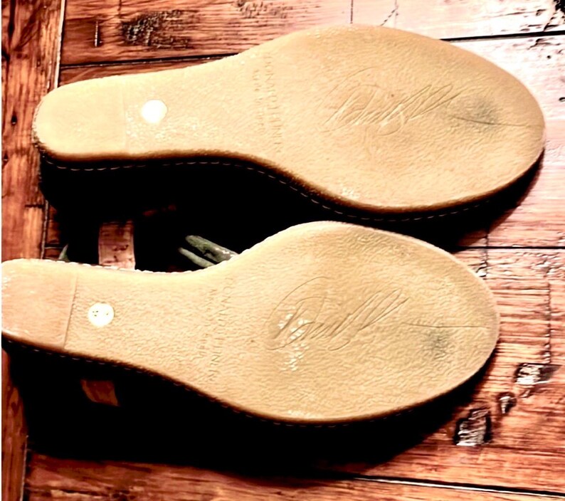 May include: Pair of wedge sandals with light brown soles and a stitched edge. The soles have a textured surface with a signature and the words "McCartney" and "Made in Italy" printed on them. The sandals are on a wooden surface.
