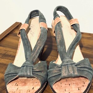 May include: A pair of open-toe wedge sandals. The sandals feature a teal fabric with a bow detail on the toe strap and a slingback strap. The footbed is made of cork, and the sole has a white stitched border. The heels are approximately 3 inches high.