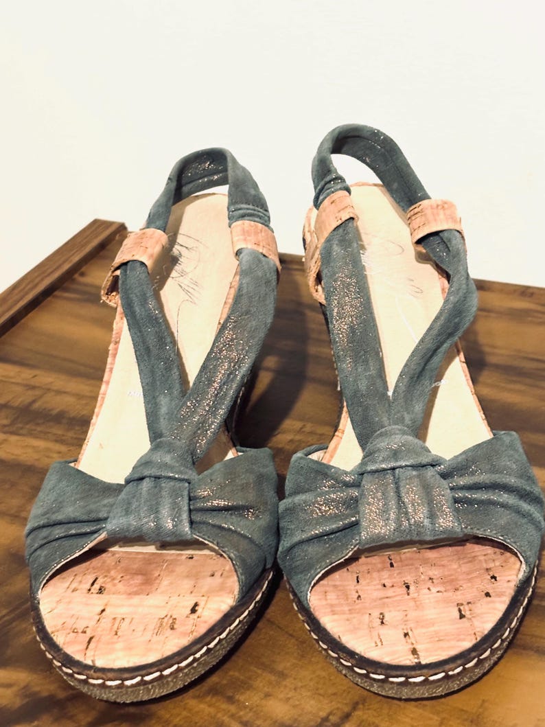 May include: A pair of open-toe wedge sandals. The sandals feature a teal fabric with a bow detail on the toe strap and a slingback strap. The footbed is made of cork, and the sole has a white stitched border. The heels are approximately 3 inches high.