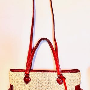 May include: A cream-coloured tote bag with red leather accents. The bag features a woven texture, red handles, and heart-shaped details. Additional red patterned pockets are on the sides. The bag has a long, adjustable strap for shoulder or crossbody wear.