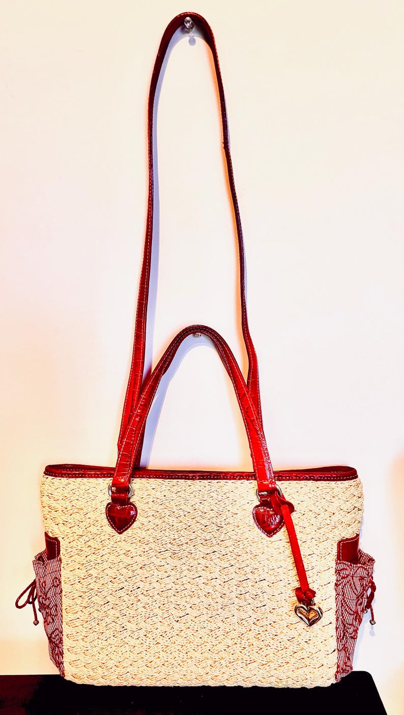 May include: A cream-coloured tote bag with red leather accents. The bag features a woven texture, red handles, and heart-shaped details. Additional red patterned pockets are on the sides. The bag has a long, adjustable strap for shoulder or crossbody wear.