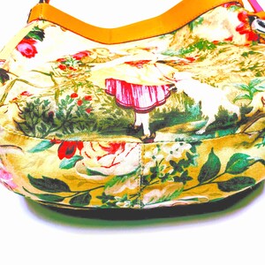 以下が含まれることがあります： 黄色い花柄のハンドバッグ、タン色の革ストラップ付き。 バッグは、バラ、緑、赤いドレスを着た人物のカラフルなプリントが特徴です。 ストラップはオレンジとピンクです。