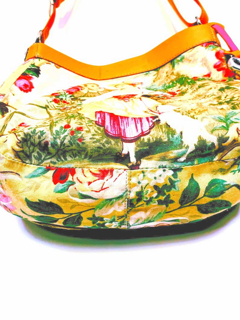 以下が含まれることがあります： 黄色い花柄のハンドバッグ、タン色の革ストラップ付き。 バッグは、バラ、緑、赤いドレスを着た人物のカラフルなプリントが特徴です。 ストラップはオレンジとピンクです。