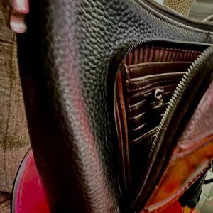 以下が含まれることがあります： 黒い革のクロスボディバッグ。表面は質感があり、バッグは開いており、茶色の内側には複数のコンパートメントとジッパーが付いています。バッグは新品のようです。